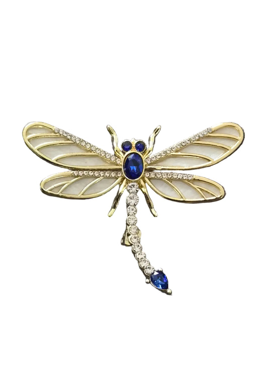 A gold dragonfly brooch with cream-colored wings, a blue gemstone body, and a tail adorned with clear crystals and a blue stone.